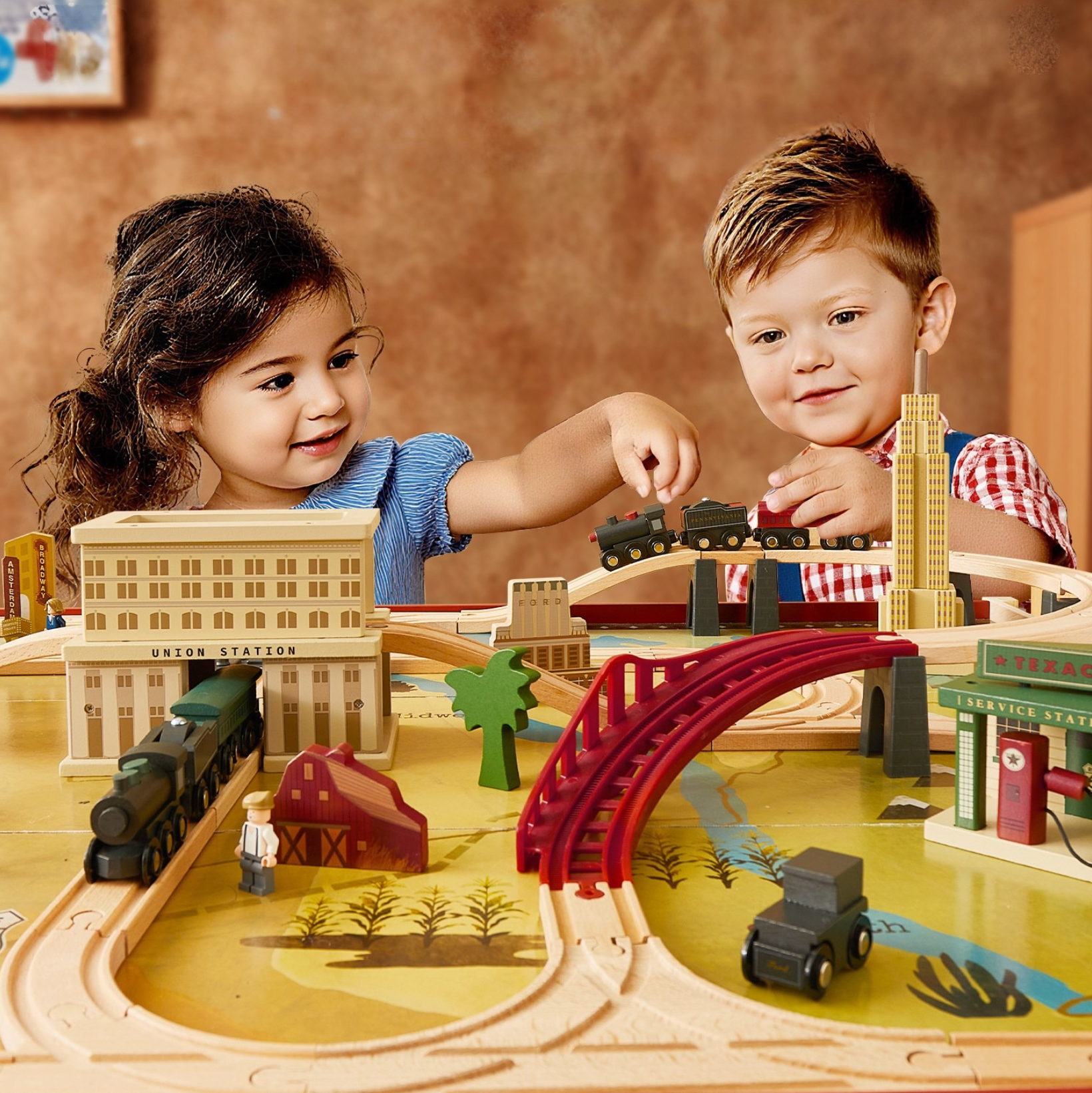A little girl wearing blue clothes and a little boy wearing a red and white checkered shirt are playing together at a wooden train track table.
