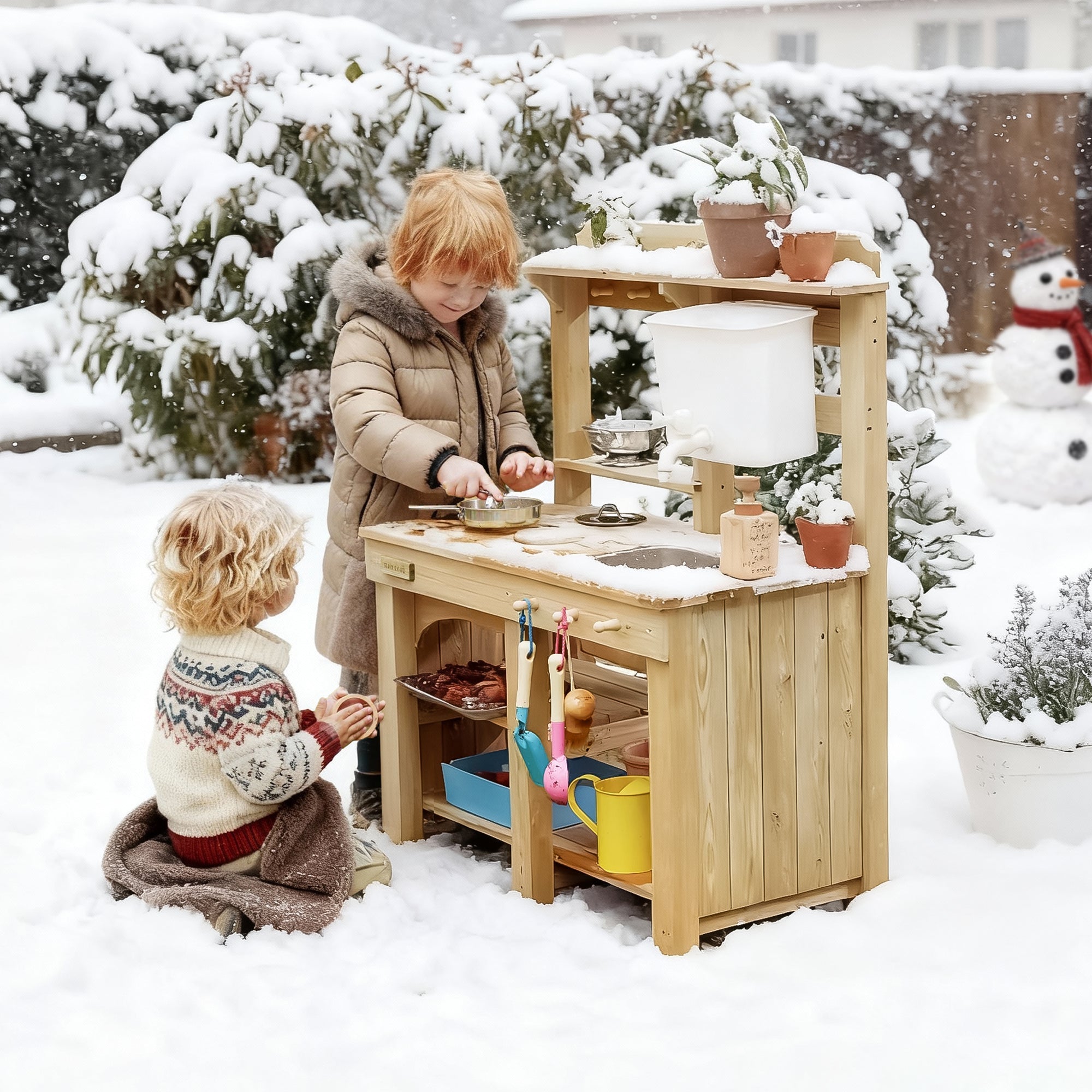 Two boys, dressed in thick winter coats, are happily playing with their outdoor play kitchen and a variety of kitchen accessories in the snowy winter outdoors.