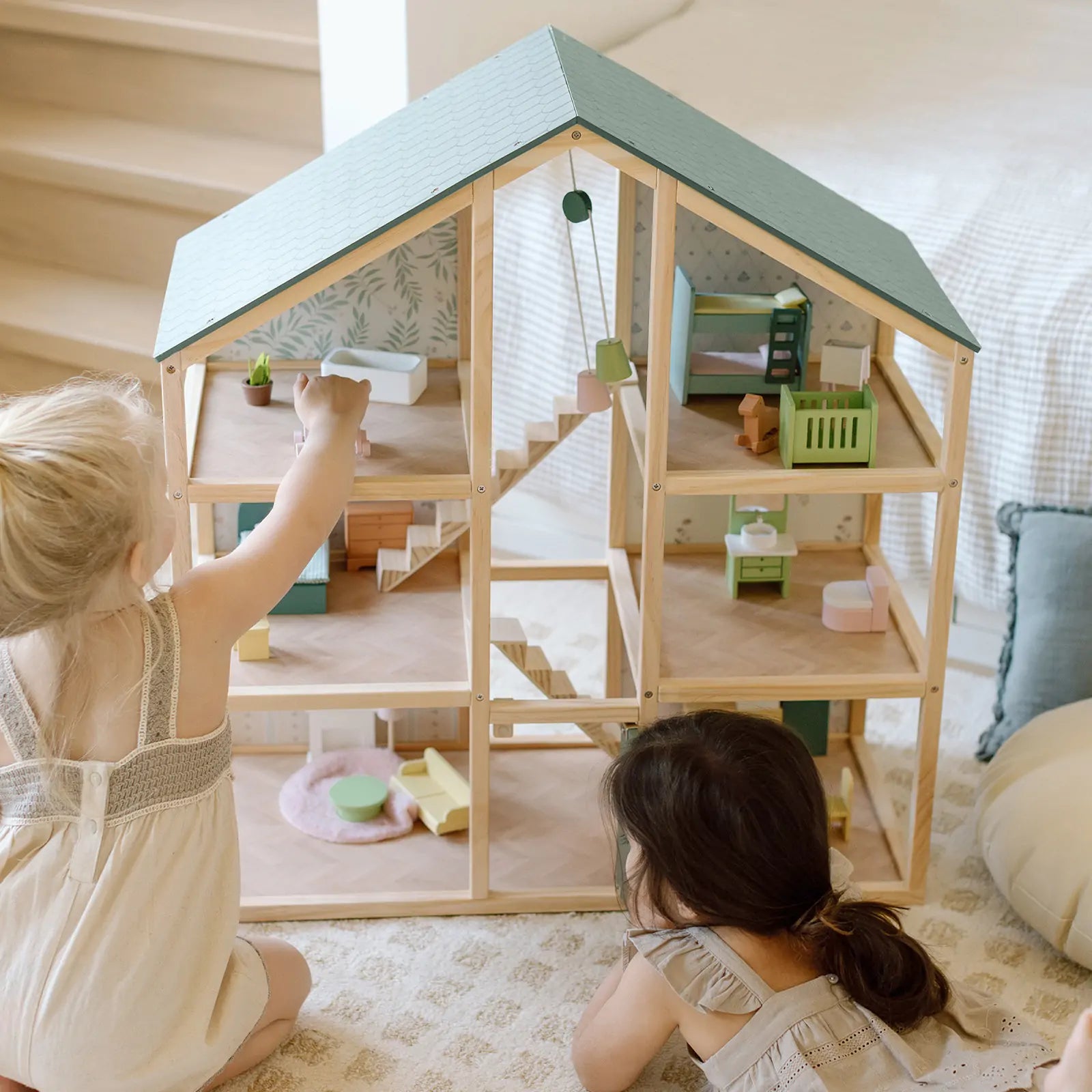 A blonde little girl and a dark-haired little girl lying on the floor playing with a green wooden dollhouse.
