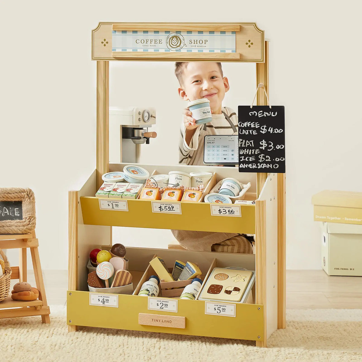 A young child smiles while playing at the Tiny Land® Farm-to-Table Gift Set—Light & Sound Kitchen & Market Stall, featuring play food, a chalkboard menu, toy coffee cups, and labeled price tags.