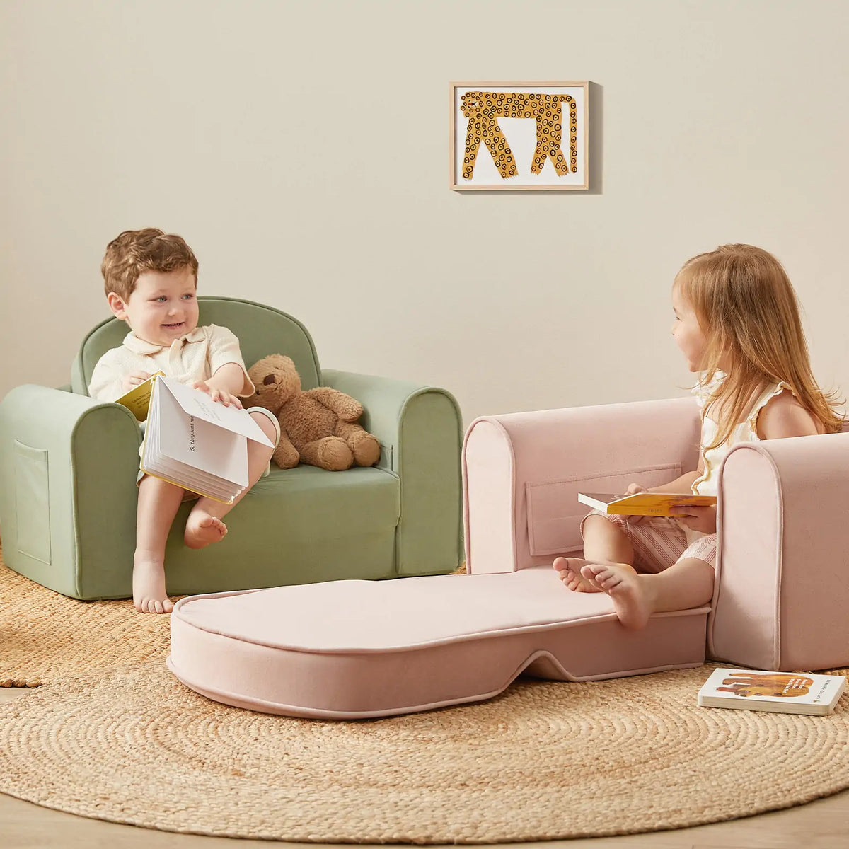 Two young children read books on pastel foldable sofas. The girl relaxes on a partially unfolded Tiny Land® GentleSnug Kid Chair - Sage, placed on a round woven rug. A plush bear and a framed animal print decorate the cozy scene.