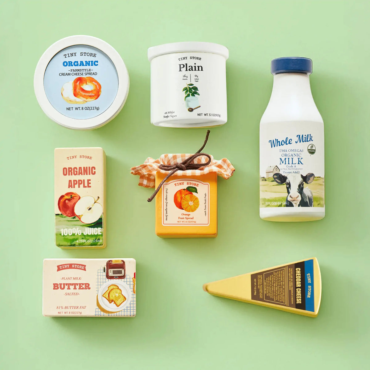 Tiny Land Play Food’s Tiny Land® Organic Dairy Pack, featuring milk, cheese, butter, apple juice, apple jam, and cream cheese—plus a wooden cutting fruit toy set—arranged on a light green background with colorful vintage-style labels.