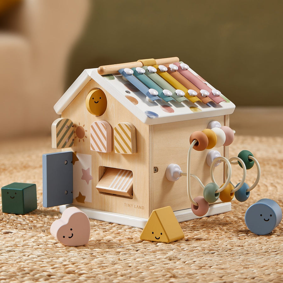 The Tiny Land Cozy Learning Playset—Playmat + Activity Cube—features pastel xylophone bars, shape sorters, a bead maze, and geometric blocks on a woven rug, making it an ideal developmental toy for toddlers.