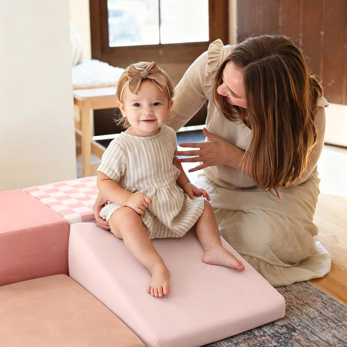 A smiling toddler in a striped dress and headband sits on Tiny Land® PlayNest Climbing Blocks, while an adult woman kneels nearby, both smiling in a cozy, sunlit room.