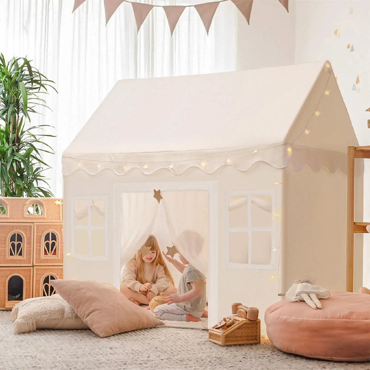 Two children enjoy the Tiny Land Bright Play & Art Set, featuring a colorful easel and a cream playhouse with lights, surrounded by pillows and toys in a bright, plant-filled room—perfect for imaginative indoor or outdoor fun|white