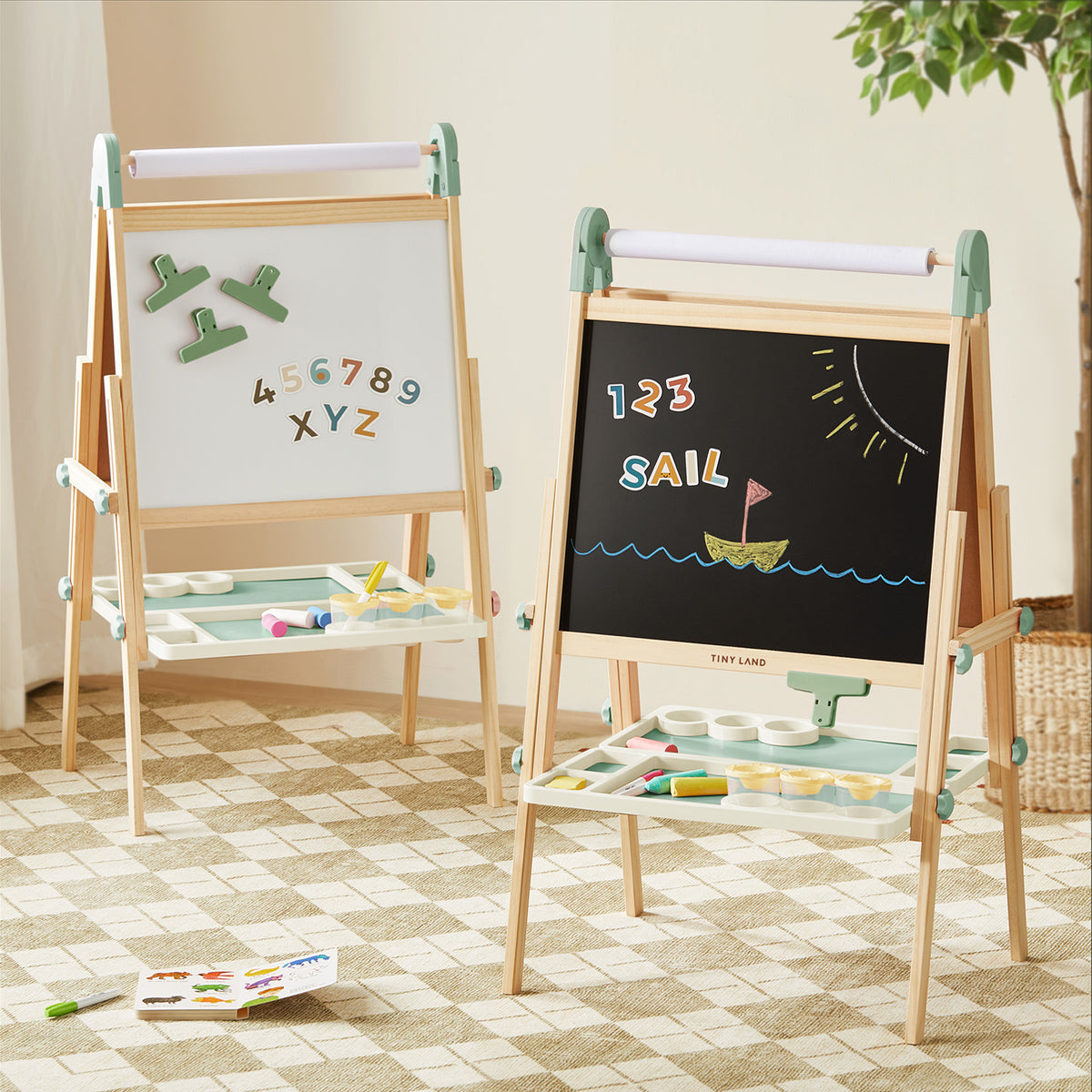 Two Tiny Land Sage Easels from the Nature-inspired Play & Art Set stand on a checkered rug—one displays a magnetized whiteboard with numbers, the other a chalkboard with a sailboat and “SAIL.” Art supplies rest on trays below, ready to inspire creativity.|green