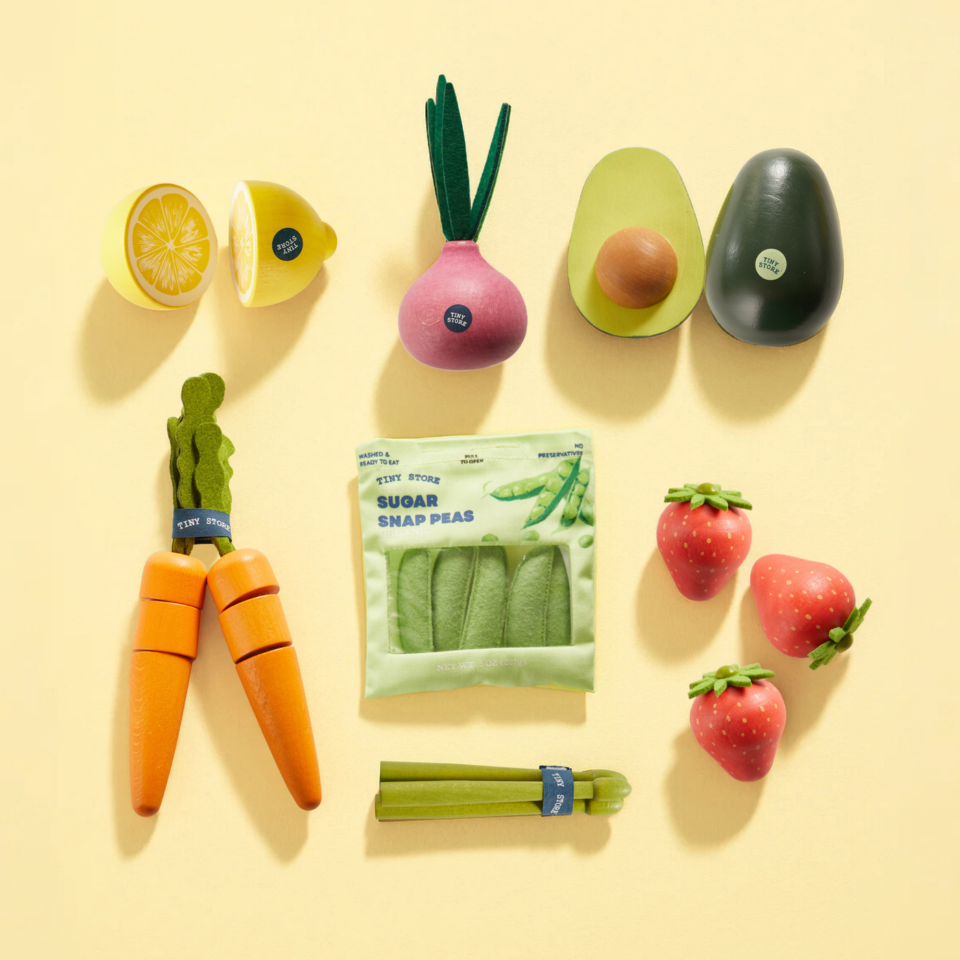 The Tiny Land® Organic Fruit & Veggie Pack by Tiny Land showcases realistic toy produce—including carrot, lemon, onion, avocado, sugar snap peas, strawberries, and celery—neatly arranged on a yellow background. These sensory toys inspire imaginative play with lifelike fruits and vegetables.