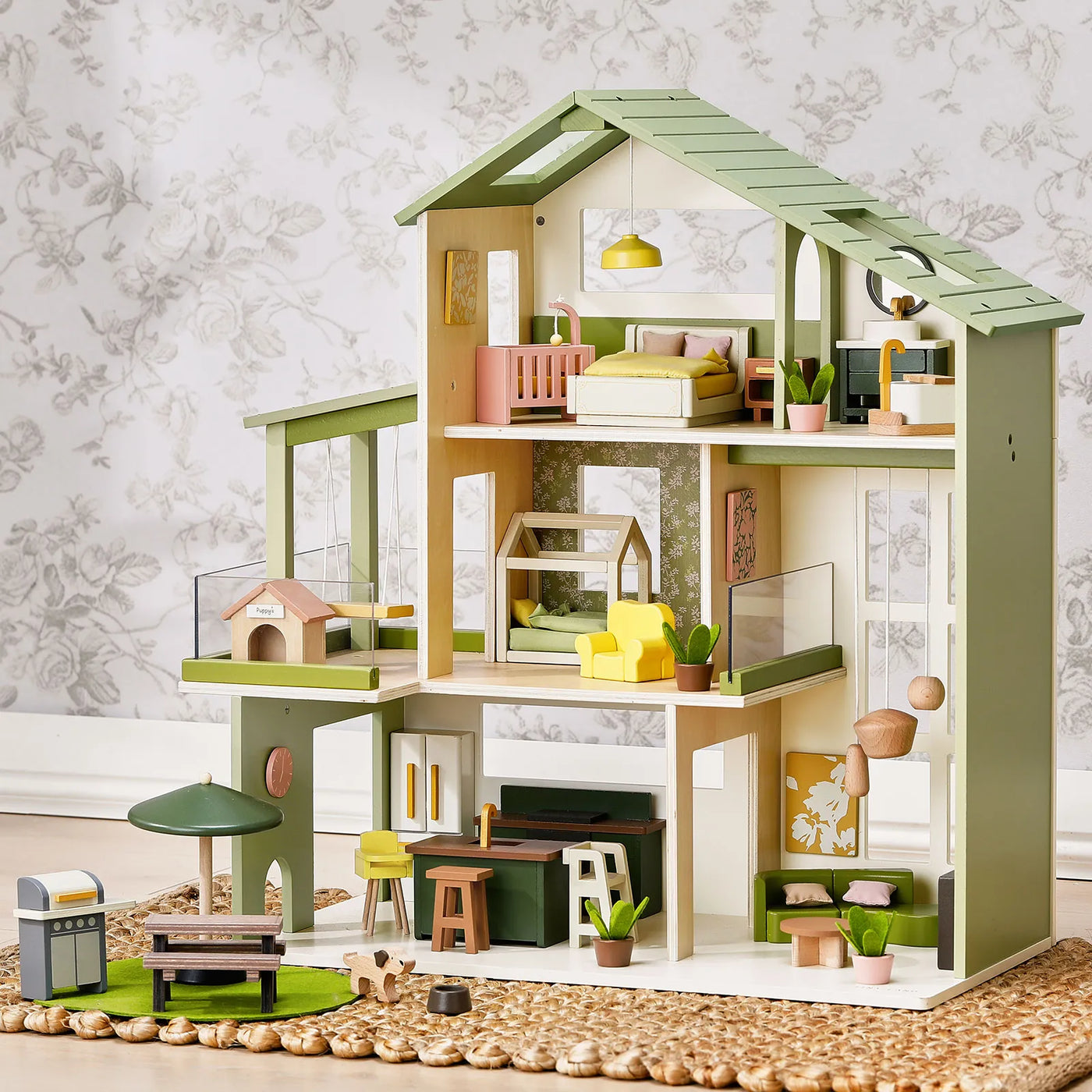 The Tiny Land® Sweetwood Edge Dollhouse - Green & Pink by Tiny Land is a modern, green-roofed wooden dollhouse featuring Montessori-inspired wooden furniture, a doghouse, grill, and patio set arranged on a woven rug, all set against a light floral wallpaper background.|Green