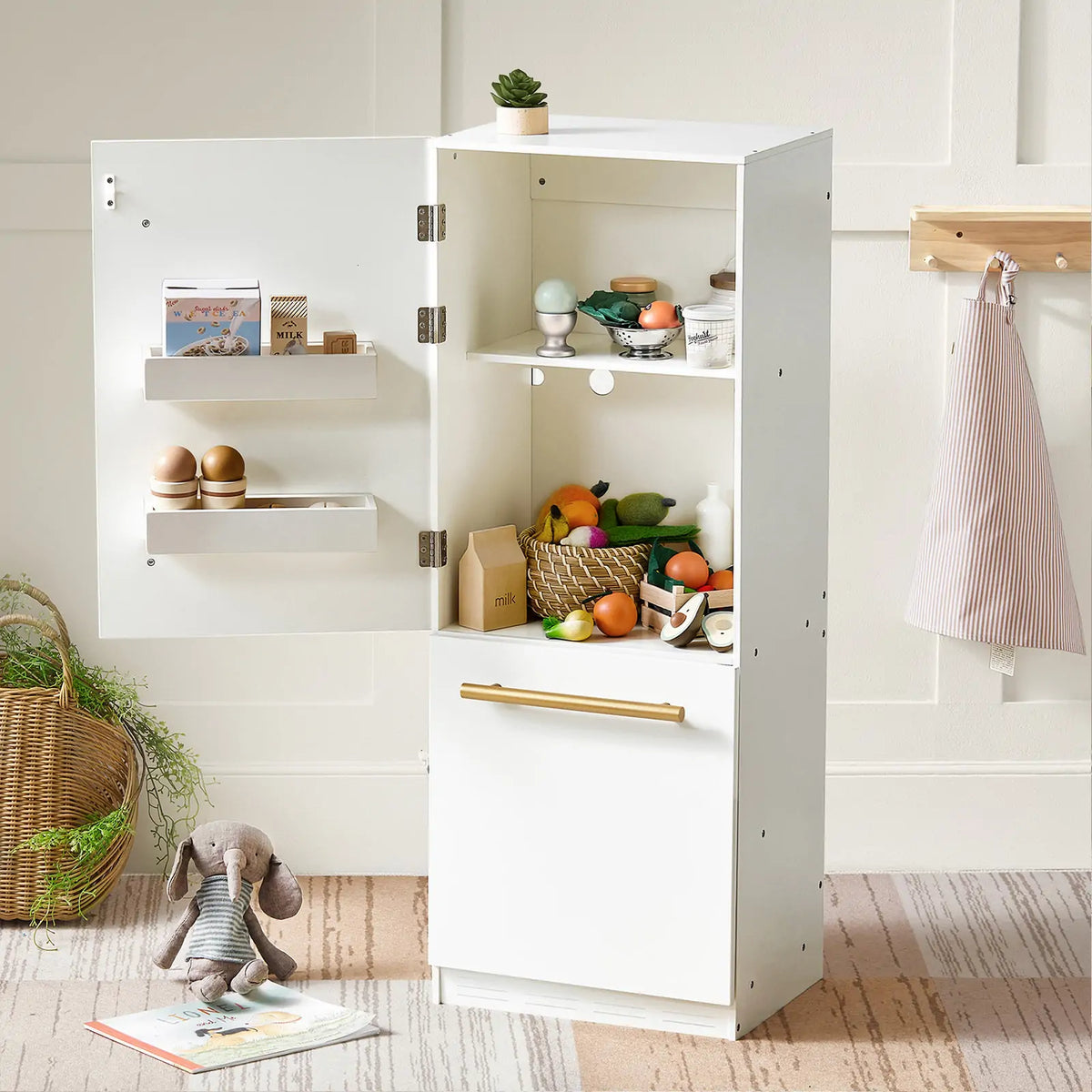 The Tiny Land® Play Kitchen Accessories – Play Kitchen Fridge is a wooden cabinet refrigerator for kids, shown open with play food, eggs, and milk inside. A plush bunny sits nearby and an apron hangs on the wall in this charming setup.