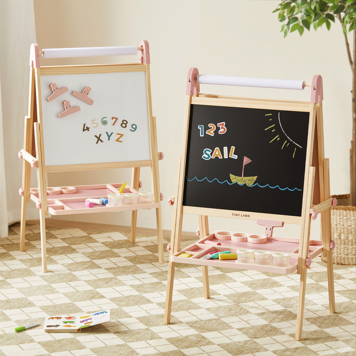 Two Tiny Land® Magnitales Easel for Kids - Pink by Tiny Land stand on a checkered rug: one displays magnetic letters and numbers, the other shows “123 SAIL” with a sailboat. Art supplies sit on trays beneath each easel.|pink