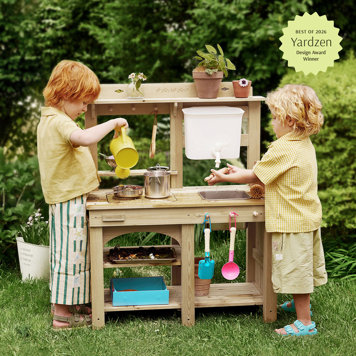 Two young children play at the Tiny Land® Mud Kitchen - DuoPlay Outdoor Play Kitchen for Kids by Tiny Land, pouring water and using toy utensils in a garden setting. Plants and kitchen items decorate the engaging sensory play setup, with a Yardzen Design Award Winner badge displayed in the top right corner.