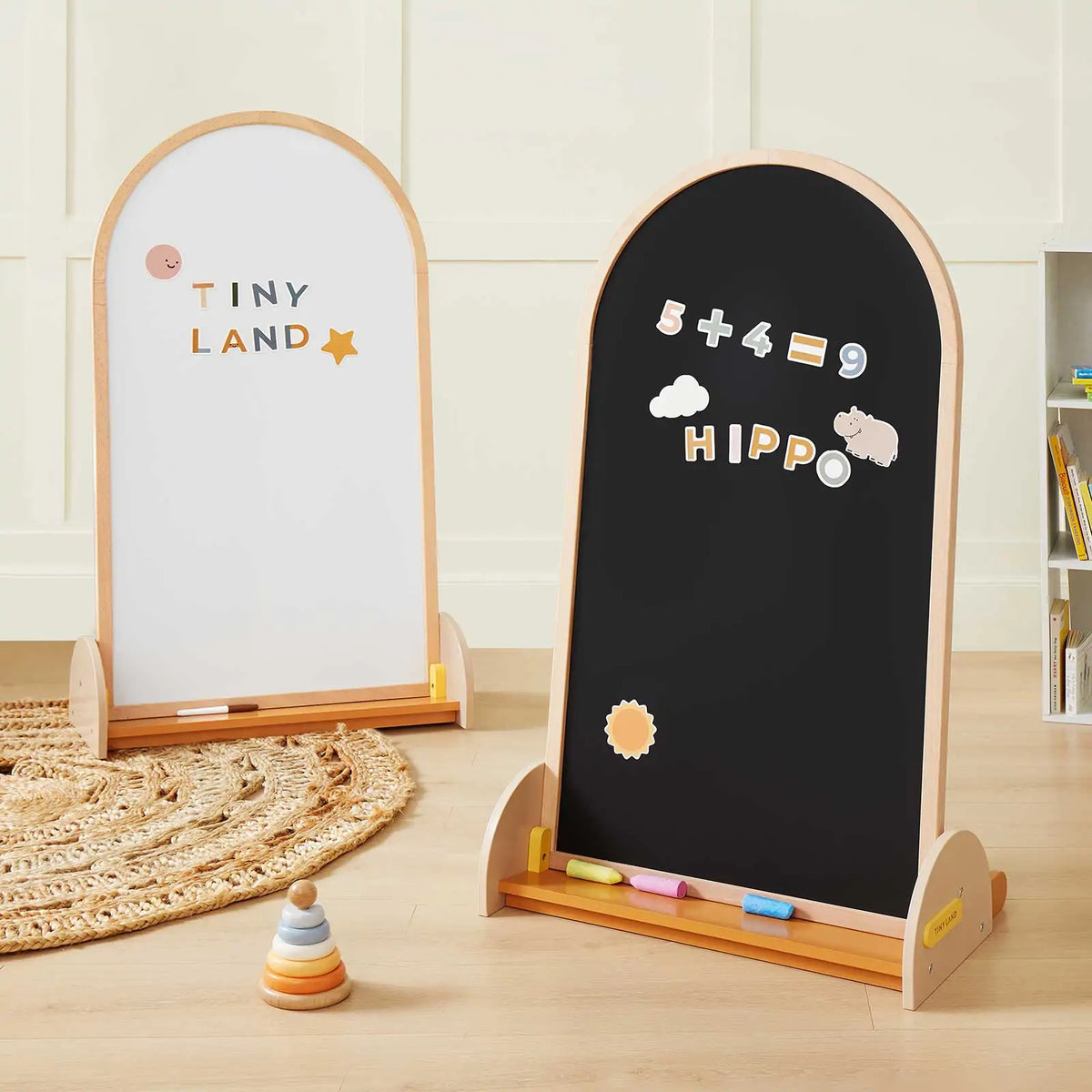 The Tiny Land® Double-Sided Arched Magnet Chalkboard from Target stands on a wooden floor; one side is a whiteboard with “TINY LAND” and a star, the other features colorful magnetic numbers, letters, and magnets. Chalk pieces and toys are nearby.