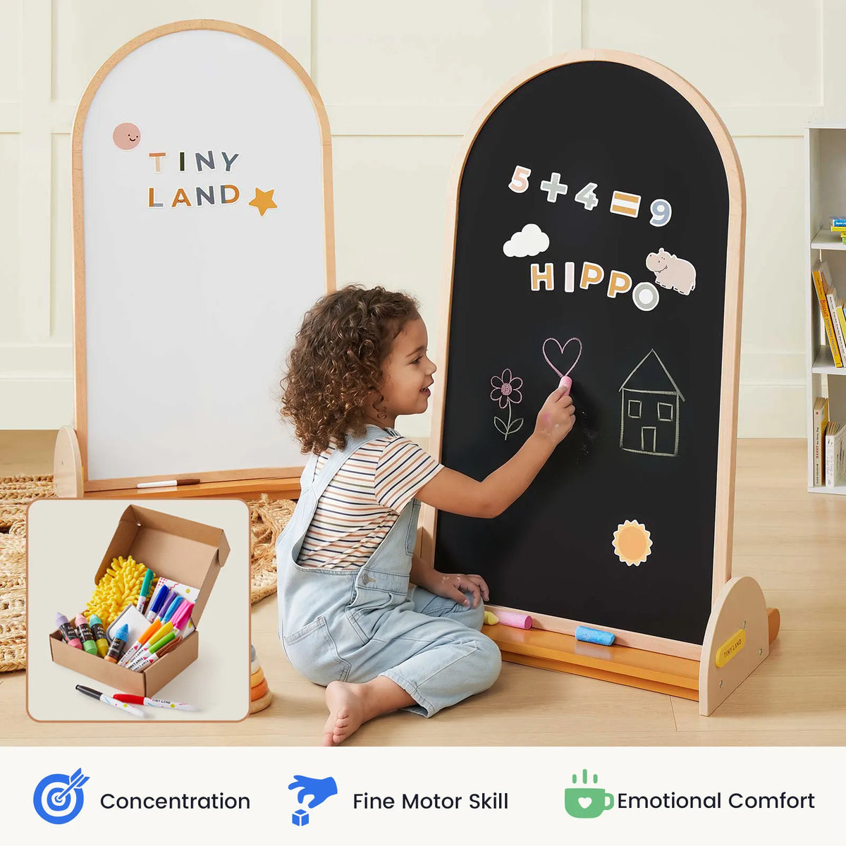 A young child draws on the Tiny Land® Double-Sided Arched Magnet Chalkboard & Art Kit using colorful chalk. Nearby, another magnetic easel from Tiny Land displays magnets spelling out "TINY LAND." In the corner, a box of art supplies and a kids' art accessory kit can be seen. Icons below highlight concentration, motor skills, and comfort.