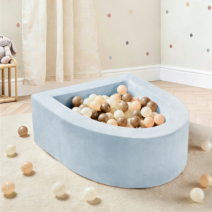 The Tiny Land® PlayNest Ball Pit with 200 Ocean Balls by Tiny Land, featuring a light blue, soft foam design and filled with pastel and neutral-colored ocean balls, sits invitingly in a nursery with wooden floors, polka dot walls, and flowing curtains. A few balls are scattered on the floor, creating a delightful play space for toddlers.|Blue