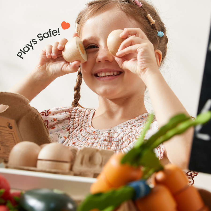 A smiling child holds two eggs over her eyes, with Tiny Land® Organic Play Food Collection and toy vegetables in the foreground. A heart and the words “Plays Safe!” appear beside her, inspiring healthy eating habits through Tiny Land Play Food kitchen toys.