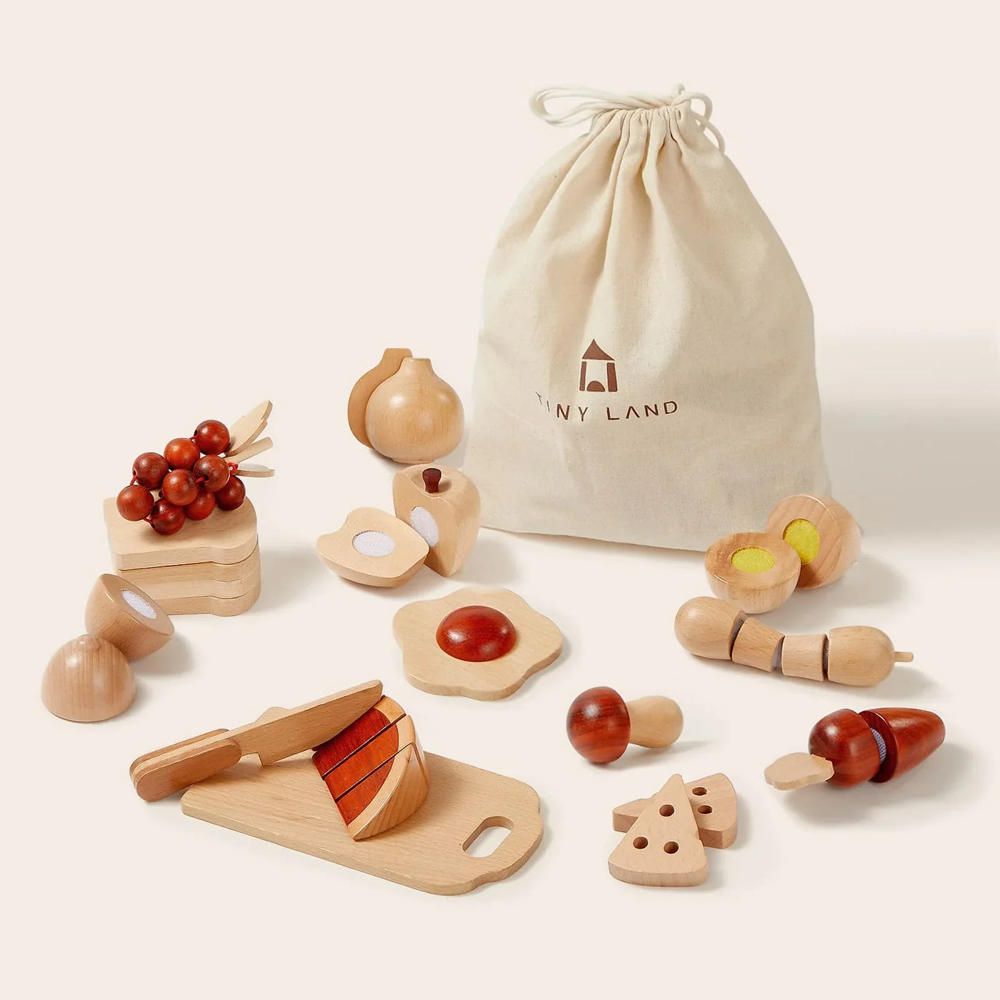 Tiny Land Play Food’s Wooden Cut and Play Food Toy Set features play kitchen accessories—fruits, veggies, cheese, eggs, a knife, and cutting board—displayed before a beige “Tiny Land” drawstring bag.