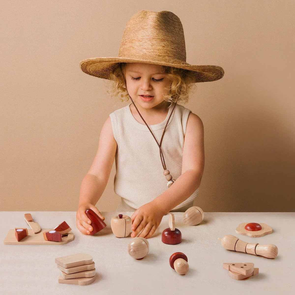 A young child wearing a wide-brimmed straw hat enjoys the Tiny Land Play Food Wooden Cut and Play Food Toys—realistic play kitchen accessories—at a beige table with a matching background.