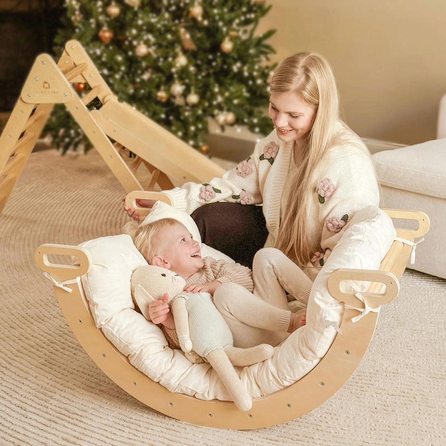 A woman and child sit in a wooden rocker with the Tiny Land® Thick Padded Play Cushion. The child holds a stuffed toy, and a decorated Christmas tree glows in the background as they smile at each other.