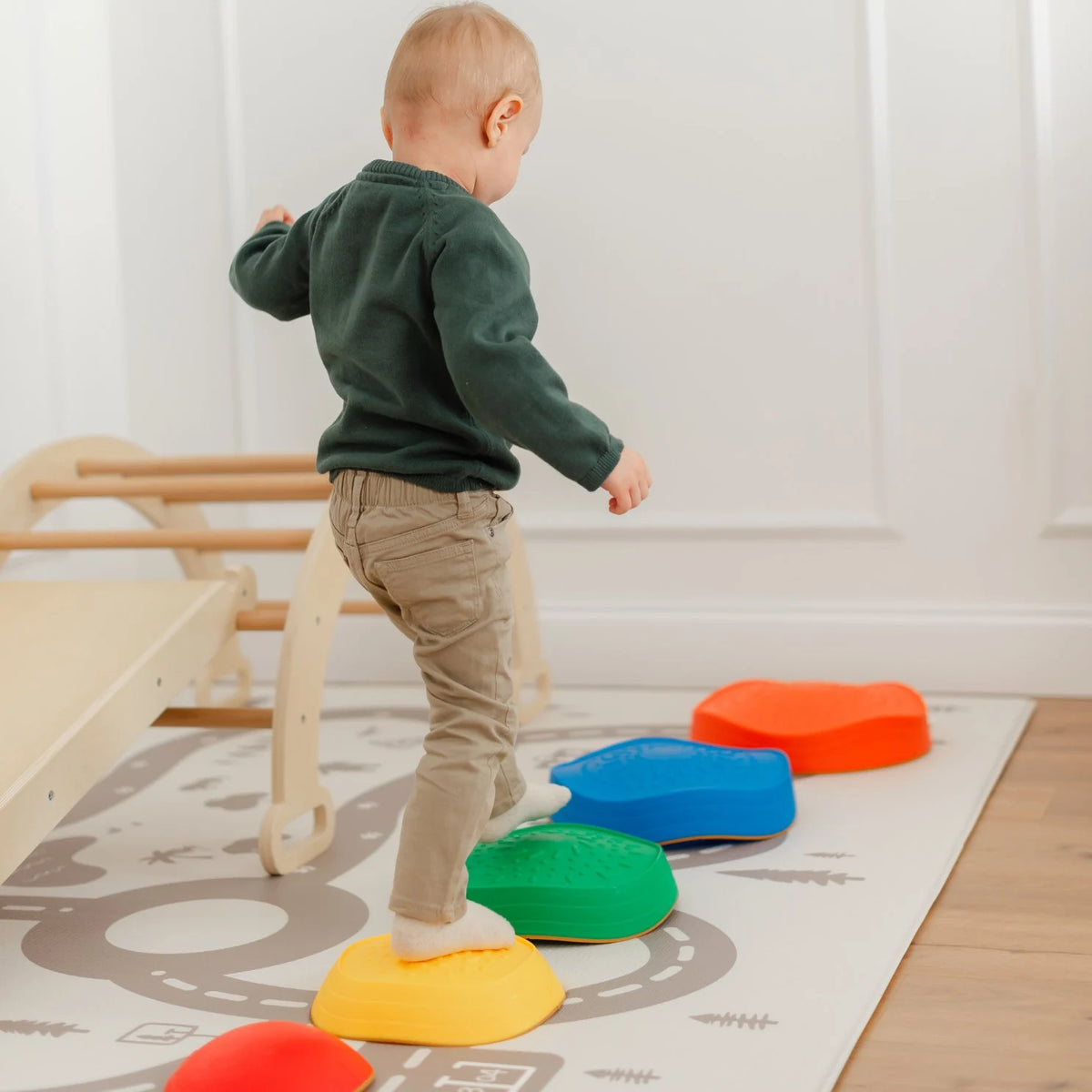 A young child in a green sweater and beige pants balances on Tiny Land's Adventure Playground Gift Set stepping stones, arranged on a play mat beside the climbing set in a bright indoor room.|Rainbow Color