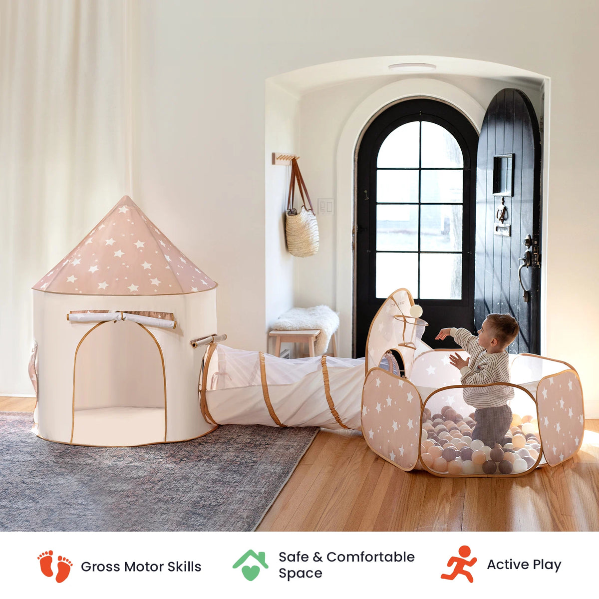A baby plays in the Tiny Land® Compact Adventure 3-in-1 Play Tunnel—featuring a star-patterned ball pit and a play tent connected by a tunnel—in a bright, cozy room filled with natural light and an arched doorway. Icons below emphasize gross motor skill development, safety, and active play from Tiny Land.|brown