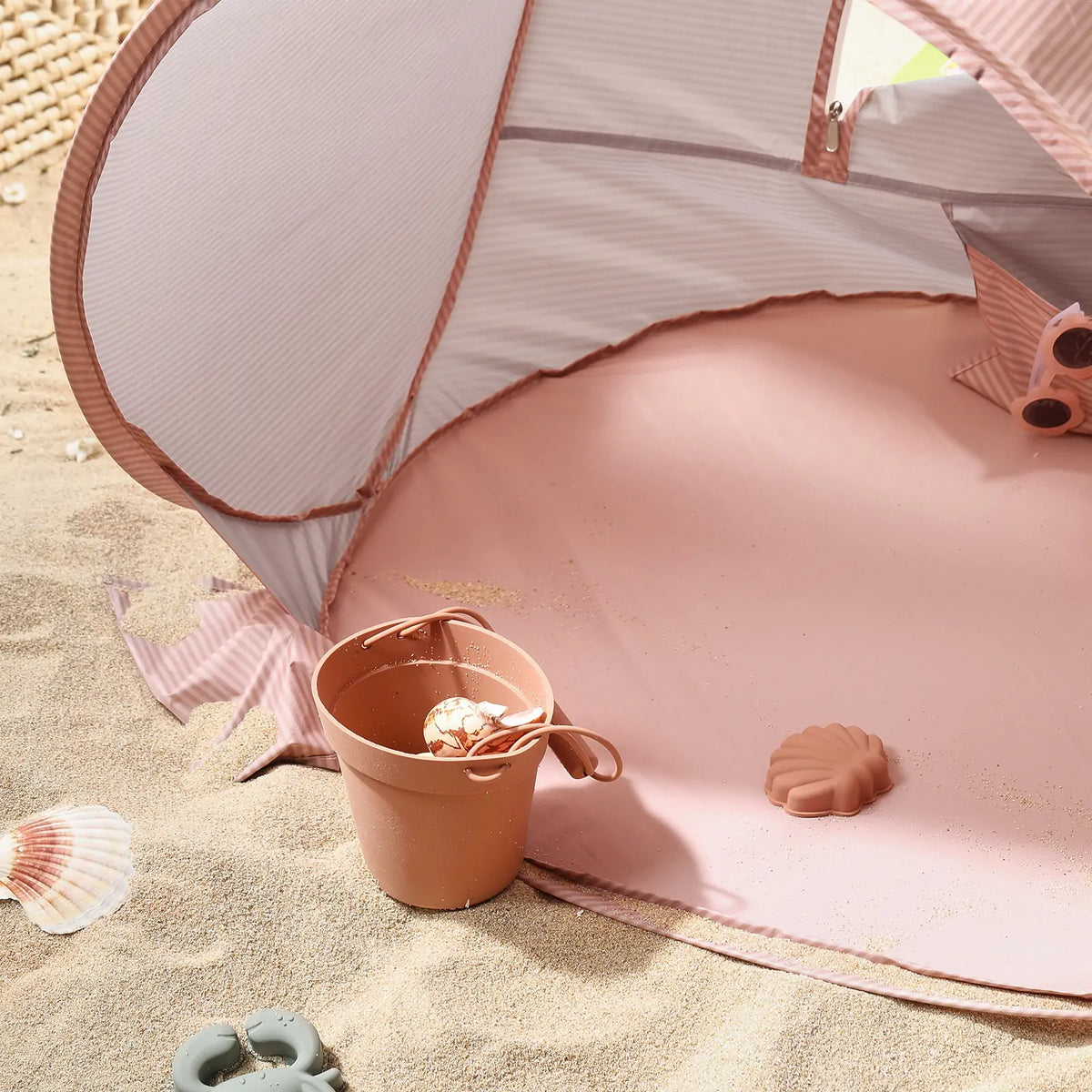 The Tiny Land® Baby Beach Tent in blush pink is set up on the sand, with a brown bucket holding a seashell and toy mold nearby. Scattered seashells complete this summery outdoor scene by Tiny Land.|Blush Pink