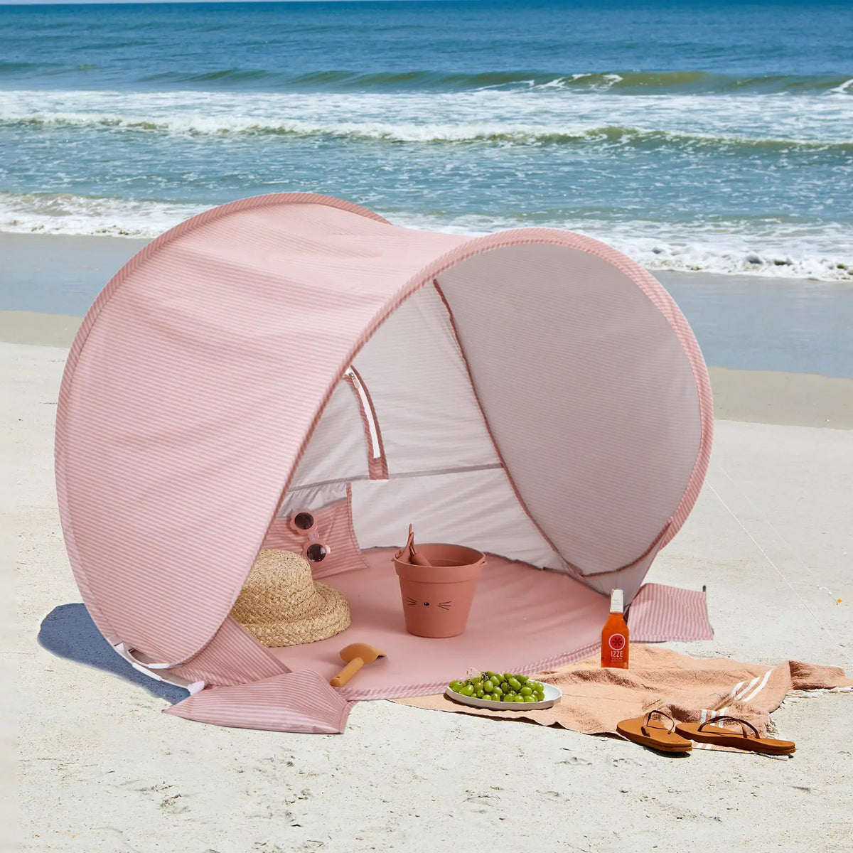 The Tiny Land® Baby Beach Tent in blush pink sits on the sand near the ocean. Inside are a straw hat, pail, sunglasses, and grapes; outside, sandals, sunscreen, and a towel await under rolling waves and a blue sky.|Blush Pink