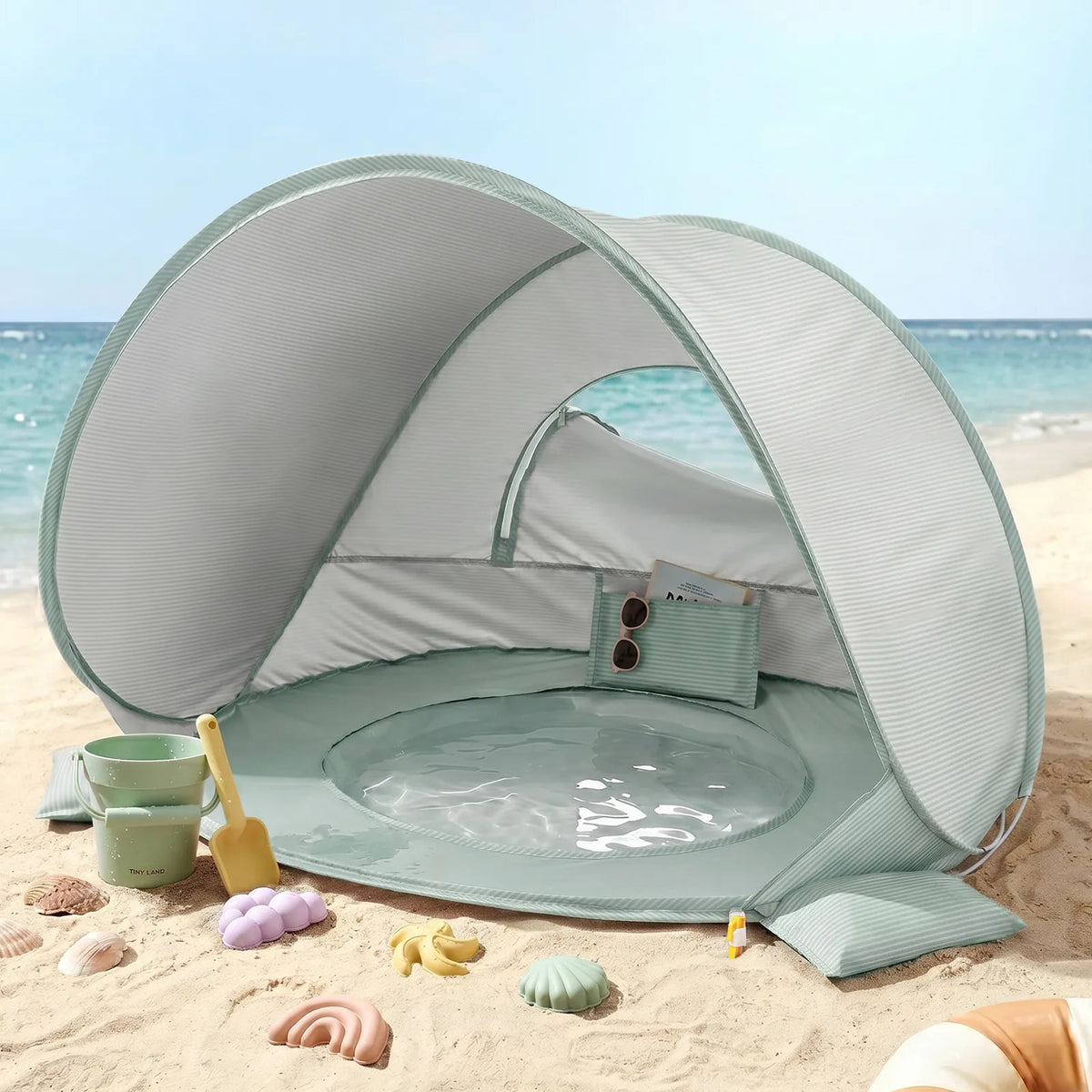 The Tiny Land® 4-in-1 Tent: Beach Tent & Sand Toy Set by Tiny Land, in a small mint-green size for toddlers, is set up on the sandy beach near the ocean. Surrounding the tent are colorful sand toys and seashells, while a pouch with sunglasses and sunscreen rests inside its built-in shallow pool.|Green