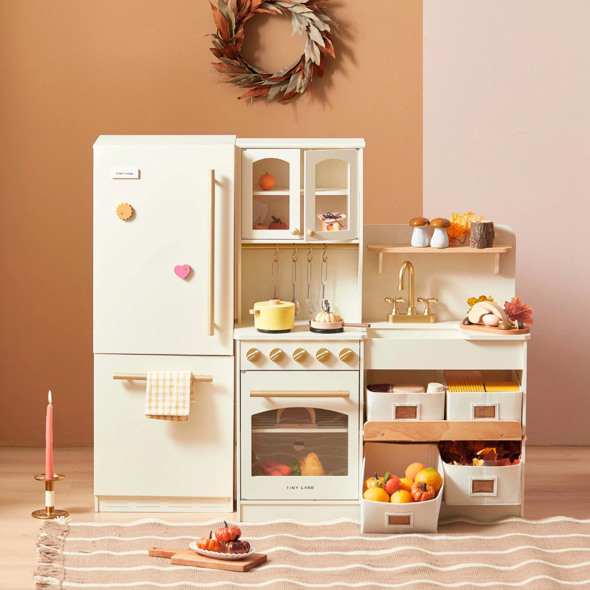 The Tiny Land Food-Filled Kitchen Gift Set—a cream kids’ kitchen with autumn decor like pumpkins, gourds, and leaves—stands by a two-tone wall topped with a leaf wreath, while a lit candle glows on the table in front.