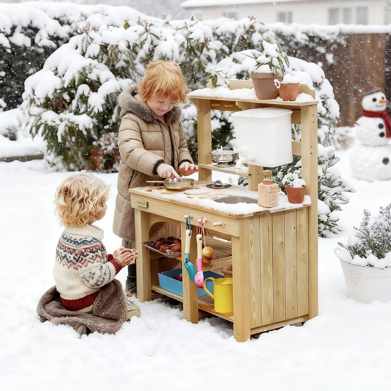 Two boys, dressed in thick winter coats, are happily playing with their outdoor play kitchen and a variety of kitchen accessories in the snowy winter outdoors.