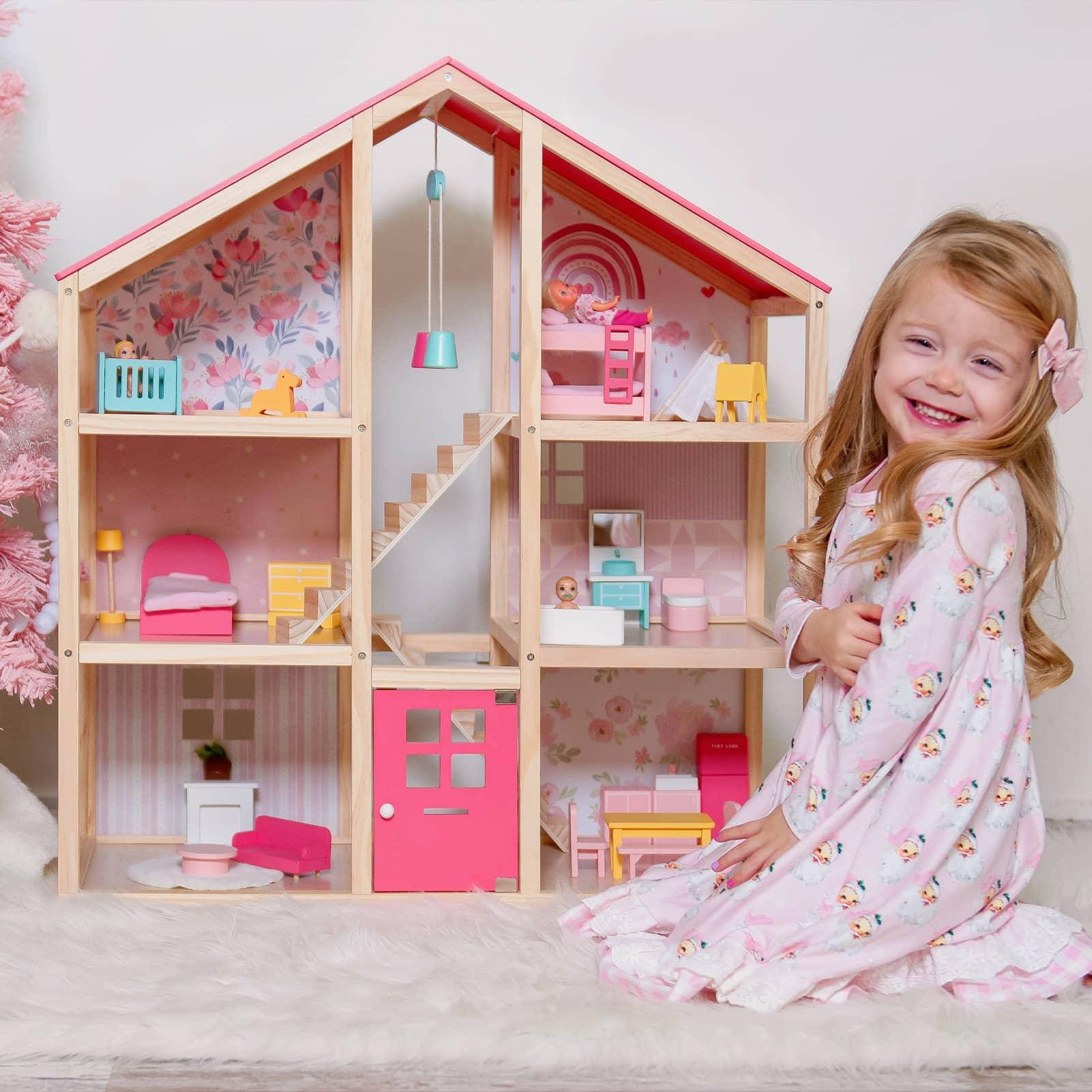 A blonde little girl sits on the floor, smiling happily beside a pink dollhouse filled with furniture.