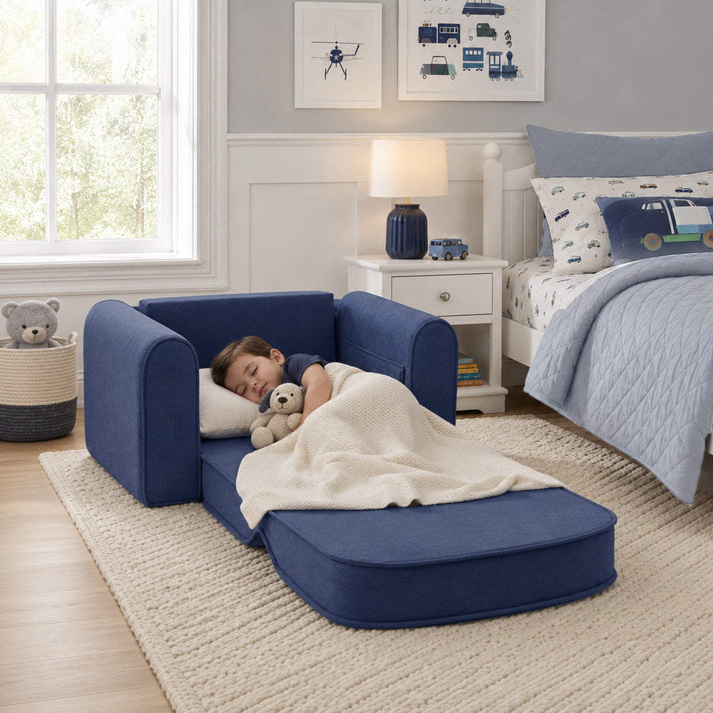 A young child sleeps on a Tiny Land® GentleSnug Kids Sofa Chair, which converts into a bed, cuddling a teddy bear and wrapped in a cream blanket in a cozy, well-lit bedroom with children's furniture and toy-themed decor.