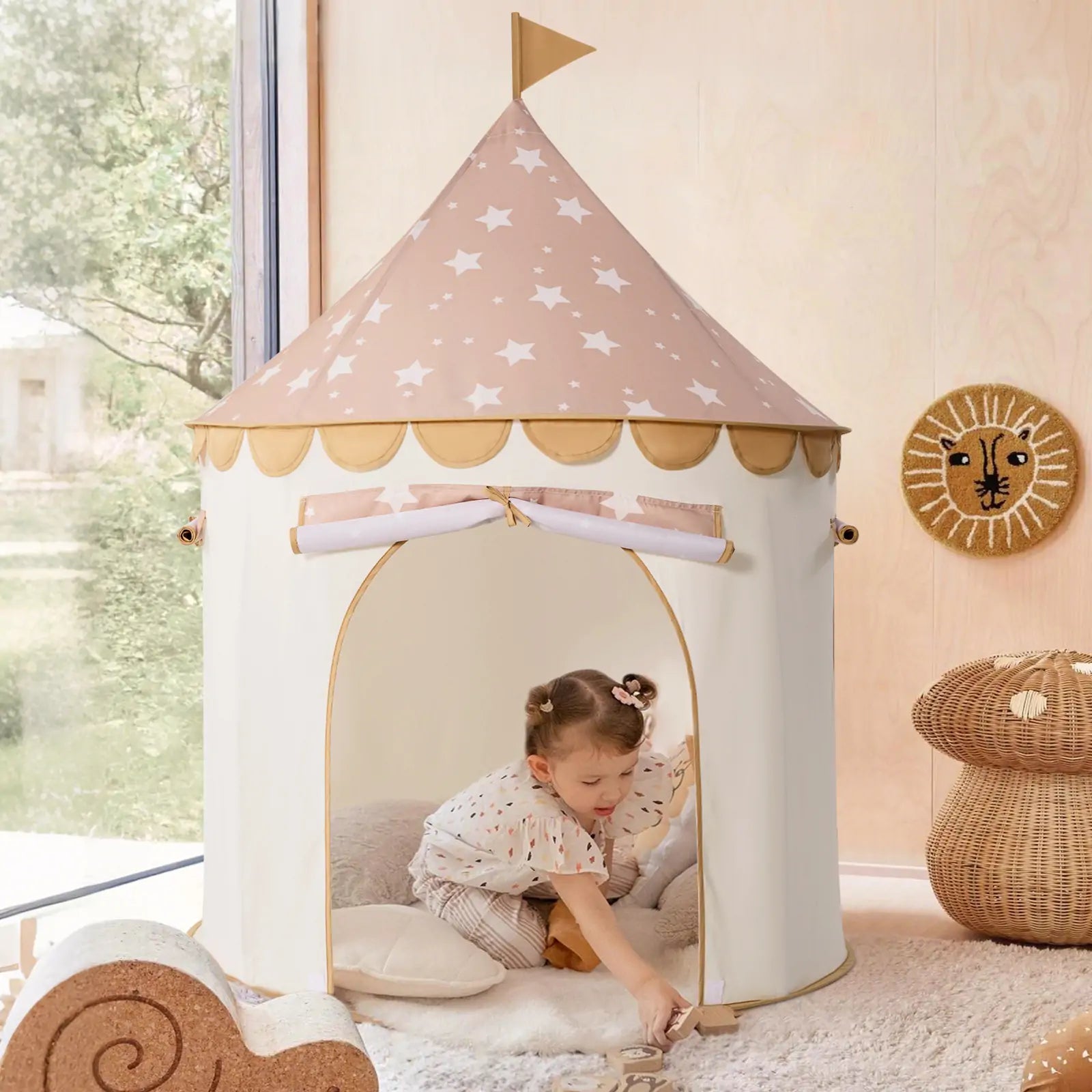 A young child enjoys the Tiny Land® Kids Tent – Indoor Tent for Toddlers, a cream play tent with a pink star-patterned roof and flag, set up indoors on a soft rug among cushions and animal-themed decor by Tiny Land.|Brown