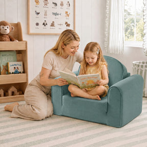 A woman kneels beside a young girl who sits in a Tiny Land® GentleSnug Kids Sofa Chair by Tiny Land, reading together in a bright, cozy children's room with toys and books neatly arranged on shelves behind them.