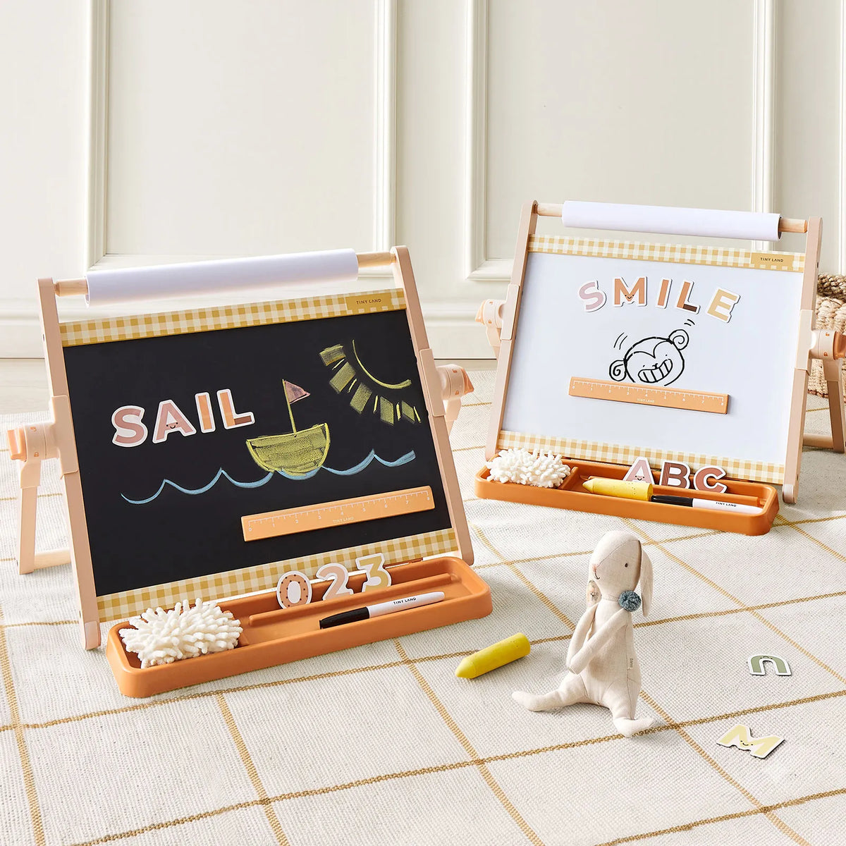 Two Tiny Land® Double-Sided Magnetic Tabletop Easels display colorful drawings and magnetic letters, setting up an inviting art station. A small plush bunny rests on the floor nearby, surrounded by chalk, erasers, and alphabet blocks in a cozy, sunlit room.|Oatmeal