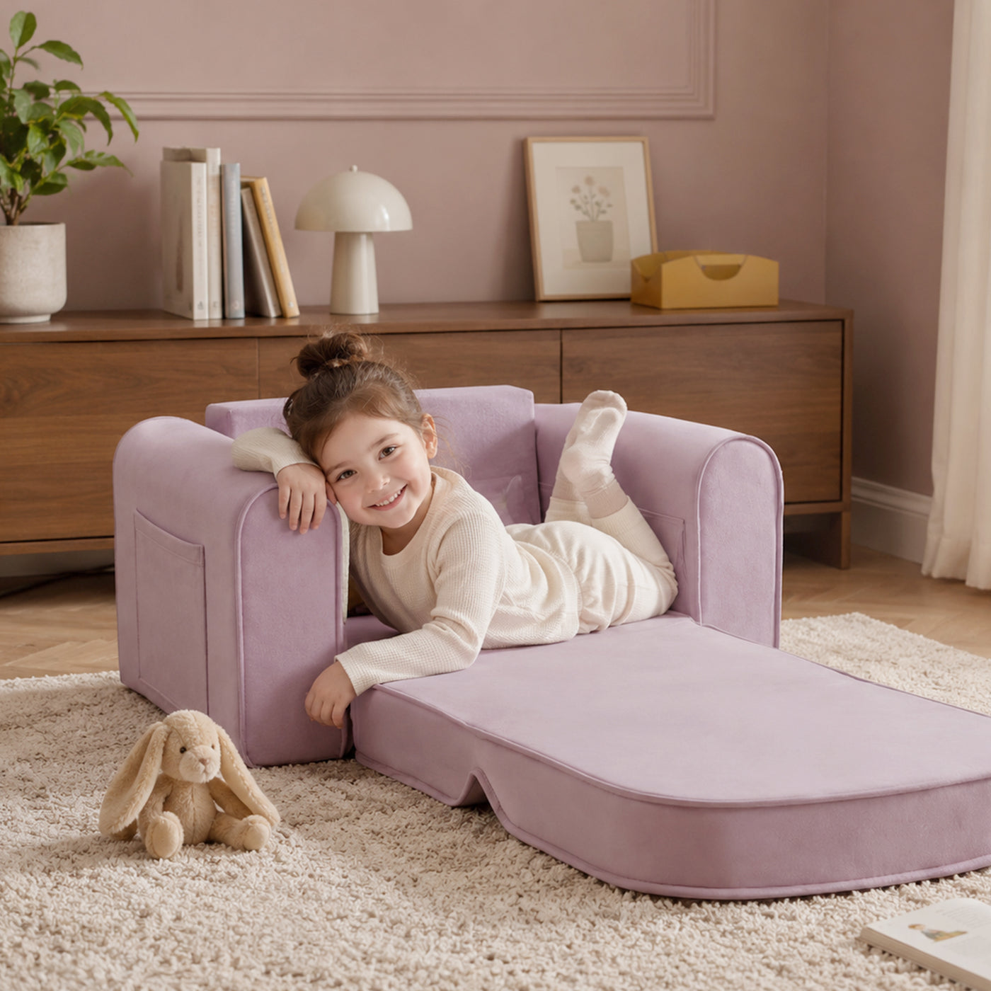 A young girl in light-colored pajamas smiles while lying on the Tiny Land® GentleSnug Kids Sofa Chair by Tiny Land in a cozy living room, with a stuffed bunny toy resting on the rug beside her.
