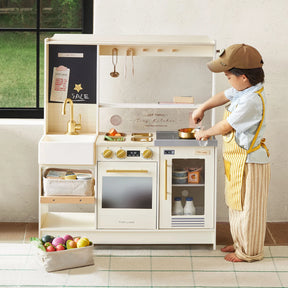 A young child in a yellow apron enjoys pretend cooking with pots and pans on the Tiny Land® BistroSwitch Play Kitchen, a white and gold set from Tiny Land. A basket of play food sits nearby as sunlight streams through the window.|White