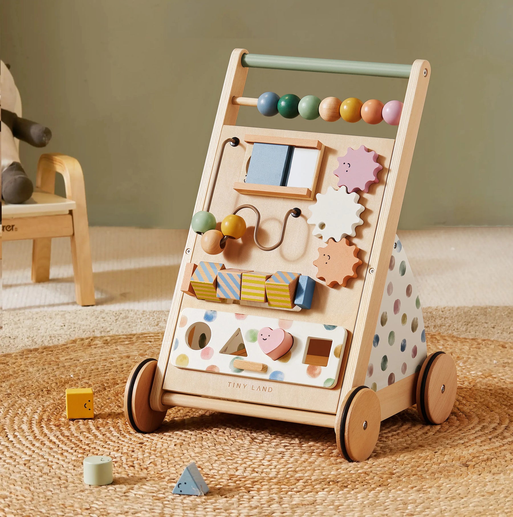 The Tiny Land Complete Development Playset — Baby Walker + Activity Cube sits on a woven rug, featuring colorful beads, gears, and shapes that encourage fine motor skills. Its sides are adorned with pastel polka dots, while nearby blocks add to the cheerful atmosphere—creating a playful space inspired by Montessori learning.