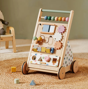 The Tiny Land Complete Development Playset — Baby Walker + Activity Cube sits on a woven rug, featuring colorful beads, gears, and shapes that encourage fine motor skills. Its sides are adorned with pastel polka dots, while nearby blocks add to the cheerful atmosphere—creating a playful space inspired by Montessori learning.