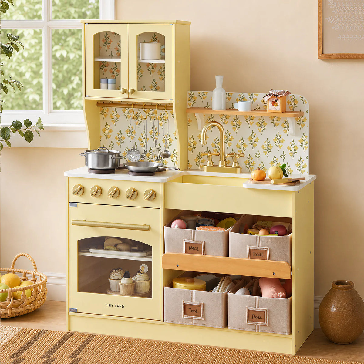 A Tiny Land® Wooden Play Kitchen with Storage for Creative Toddlers by Tiny Land, featuring a yellow finish, stove, oven, sink, shelves, and multifunctional storage bins, stands against a wall adorned with floral wallpaper. The kitchen is filled with pretend cookware, dishes, and play food, while a basket of plush food sits on the floor nearby.|Pale Yellow