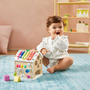 A smiling baby in a white outfit sits on a blue rug, playing with the Tiny Land Rainbow Baby Walker & Activity Cube Developmental Playset, designed to promote fine motor skills. Behind them are a pink wall, cushion, and shelves with toys and decor.