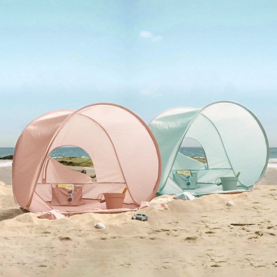 Two Tiny Land® One-Stop Beach Toy & Tent Sets—one in blush pink, one in light blue—are set up on the sand, each with a matching play set inside and ocean views, perfect for fun and UV protection.