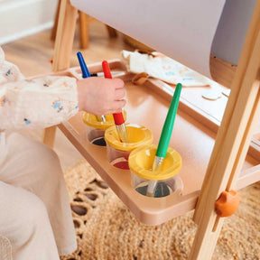 A child dips a brush into yellow paint on the Tiny Land® Caramel Magnitales Easel for Kids, with red and green paint pots nearby, a paper roll above, and a woven rug on the floor.