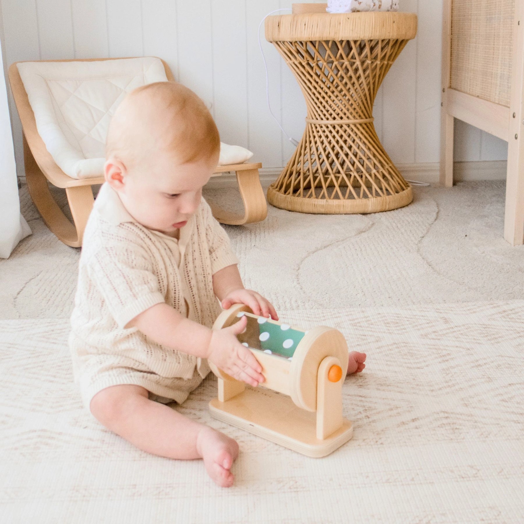 A baby sits on a light rug in a neutral-toned nursery, playing with Tiny Land® Montessori Toys for Newborns (4–6 Months) by Tiny Land. A wicker side table and small chair are in the background.
