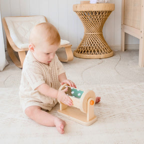 A baby sits on a light rug in a neutral-toned nursery, playing with Tiny Land® Montessori Toys for Newborns (4–6 Months) by Tiny Land. A wicker side table and small chair are in the background.