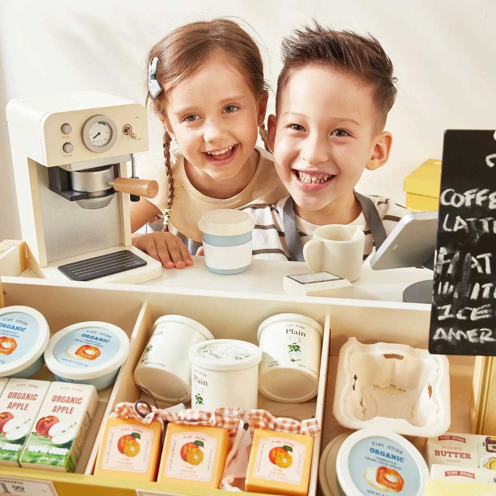 Two smiling children stand behind the Tiny Land® Farm-to-Table Gift Set—a Light & Sound Kitchen & Market Stall by Tiny Land—featuring a toy espresso machine, cash register, and pretend organic foods like yogurt, eggs, and apple juice for endless play.