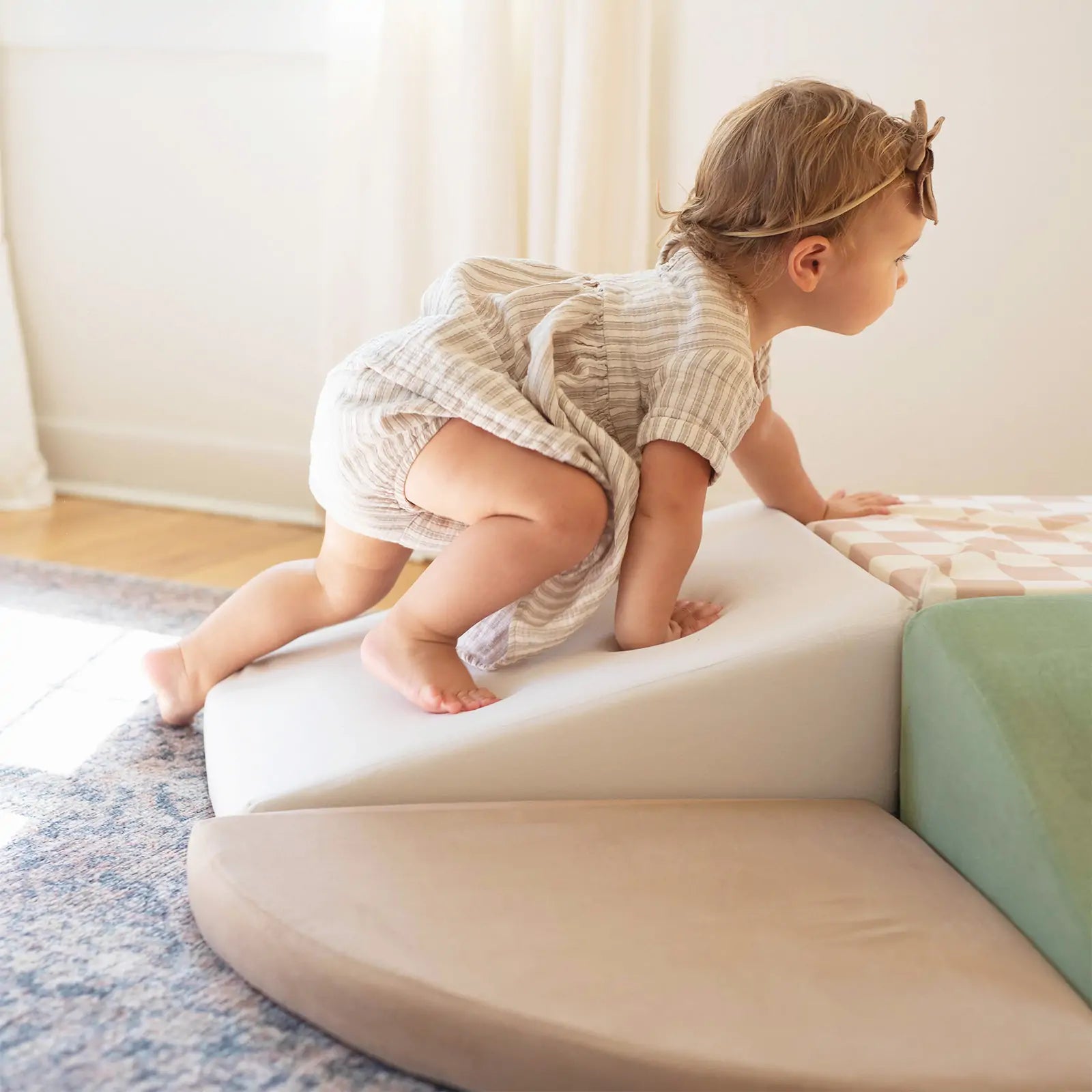 A toddler in a striped dress and headband climbs colorful blocks on the Tiny Land® PlayNest Climbing Ball Pit Set with Tunnel by Tiny Land, enjoying playtime on the soft foam structure in a bright, sunlit room with a rug.