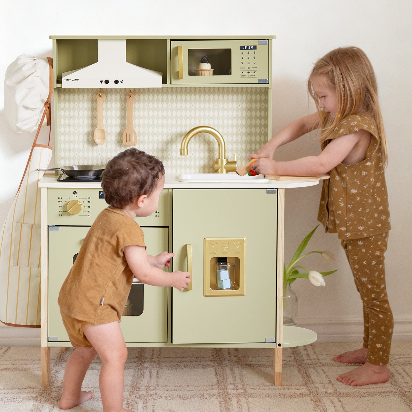 Two young children play with the Tiny Land® PopJoy Play Kitchen in green. One child stands at the sink washing toy food from the set, while the other stands nearby. Both are dressed in brown outfits. A tulip placed on a nearby shelf adds a cheerful touch to their pretend play with this delightful Tiny Land kitchen.|Sage