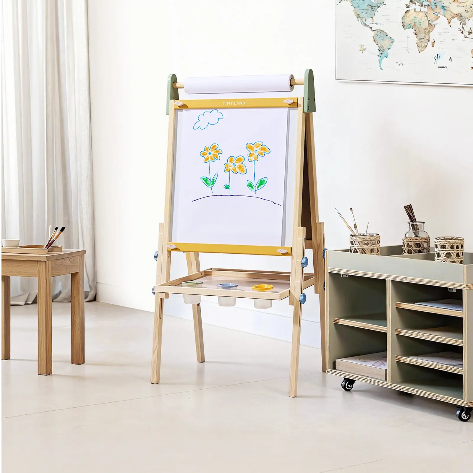 The Tiny Land® 3-in-1 Art Easel for Kids - Morandi stands in a bright room with a painting of yellow flowers and a cloud, next to art supplies, brushes on a small table, a storage cart, and a world map on the wall.|green
