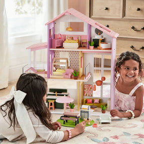 Two young girls smile and play with the Tiny Land® Sweetwood Cottage Dollhouse on a patterned rug. The colorful dollhouse from Tiny Land features multiple rooms, tiny furniture, and fun accessories in a bright room.|Pink
