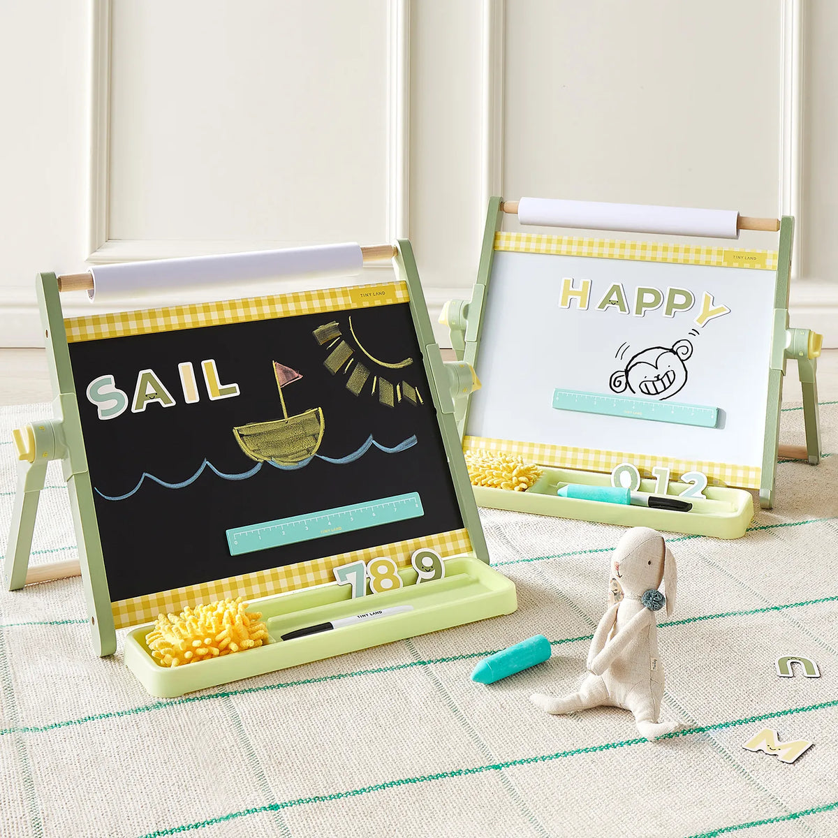 Two light green Tiny Land® Double-Sided Magnetic Tabletop Easels sit on a rug. The left adjustable easel by Tiny Land features a chalkboard displaying "SAIL" with a boat drawing, while the right side offers a magnetic whiteboard showcasing "HAPPY" and a monkey illustration. A plush bunny sits in the foreground next to scattered letters, making this the perfect kids’ art station for creativity and play.|Green